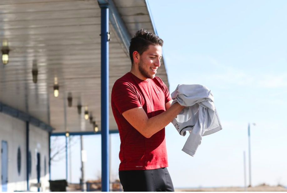 A smiling personal trainer in a red shirt holds a grey towel or shirt while standing outdoors.