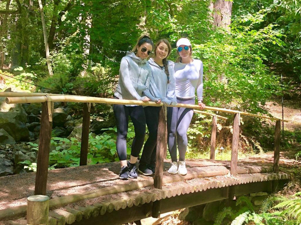 Three women in athletic wear stand on a wooden bridge in a lush, green forest, enjoying an outdoor activity.