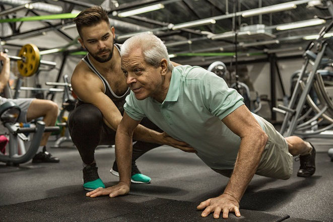 A personal trainer assists an older client with a push-up in a gym, demonstrating attentive coaching.