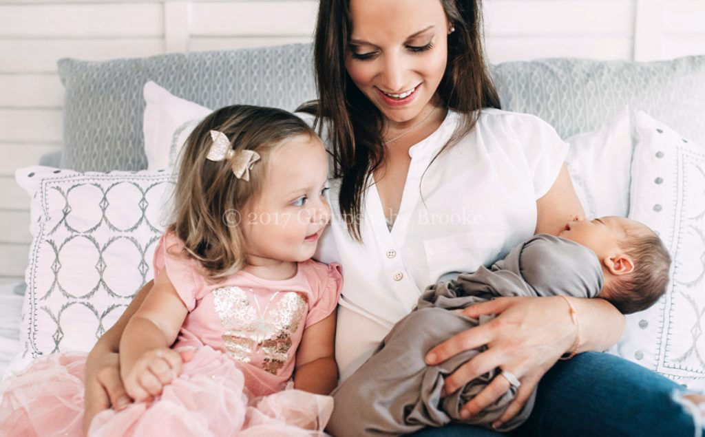 A mother smiles down at her newborn baby, held in her arms next to her young daughter in a pink dress.