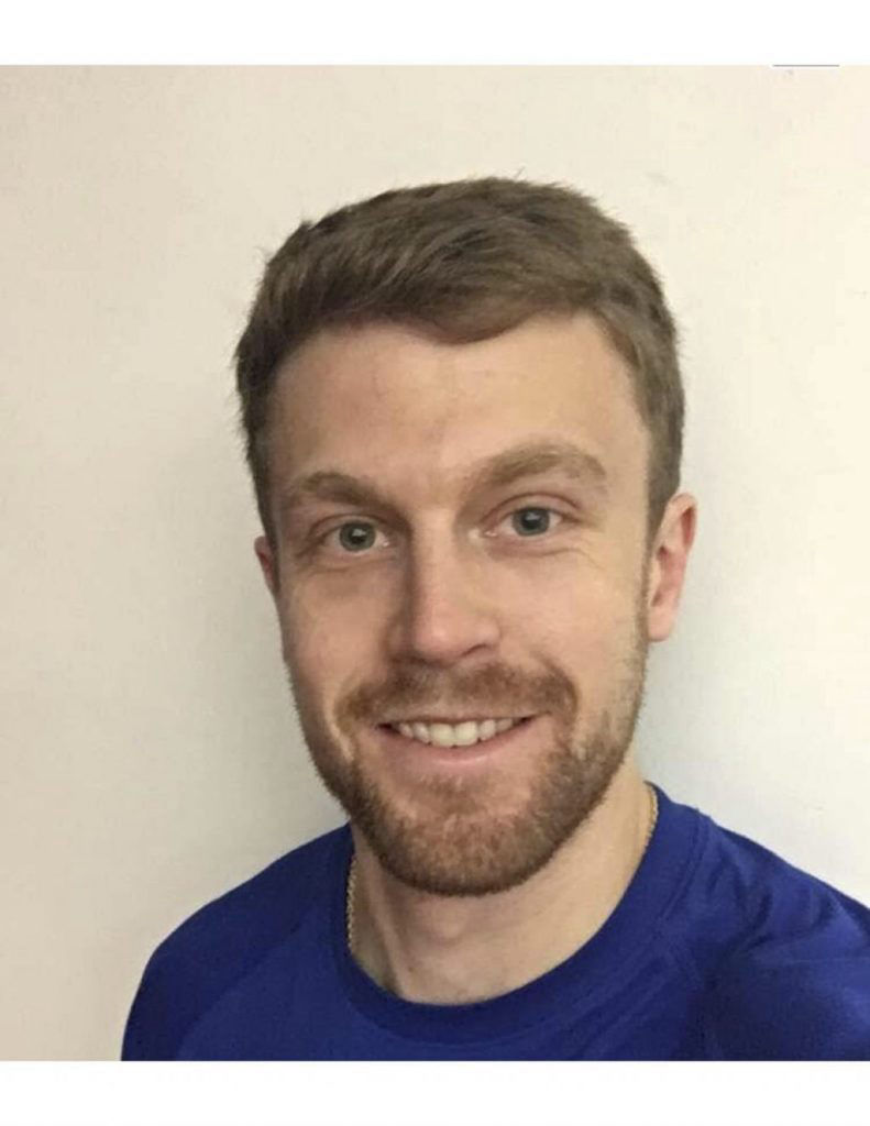 Headshot of a personal trainer with a beard, smiling slightly in a blue shirt.